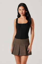 A woman wearing a sleeveless black top with a square neckline and button details down the front, paired with a high-waisted plaid skirt featuring pleats.
